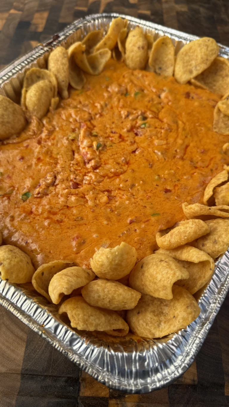 Finished smoked longaniza queso dip in aluminum pan surrounded by tortilla chips, showing smooth melted cheese with visible sausage pieces and vegetables