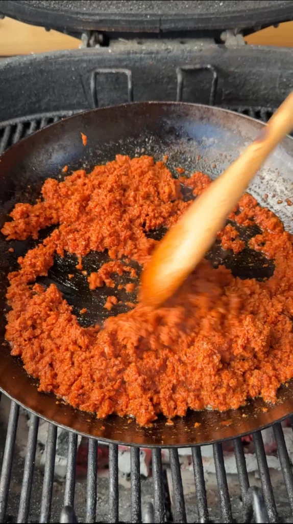 Longaniza sausage cooking in cast iron skillet on grill with wooden spoon, showing browned sausage pieces ready for queso dip recipe