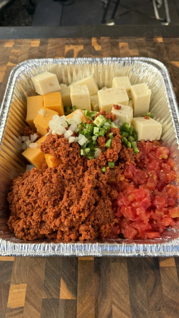 Raw ingredients for smoked longaniza queso dip arranged in aluminum pan showing cubed Velveeta, cheddar, pepper jack cheese, diced onions, jalapeños, and Rotel tomatoes