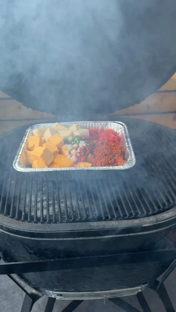 Aluminum pan of longaniza queso dip smoking on grill grates with visible smoke, showing ingredients before melting process begins