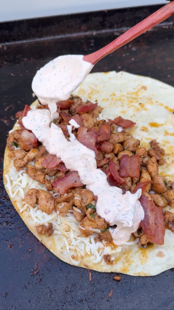 Homemade chipotle ranch sauce being drizzled from wooden spoon onto seasoned chicken and bacon mixture in large flour tortilla on black griddle