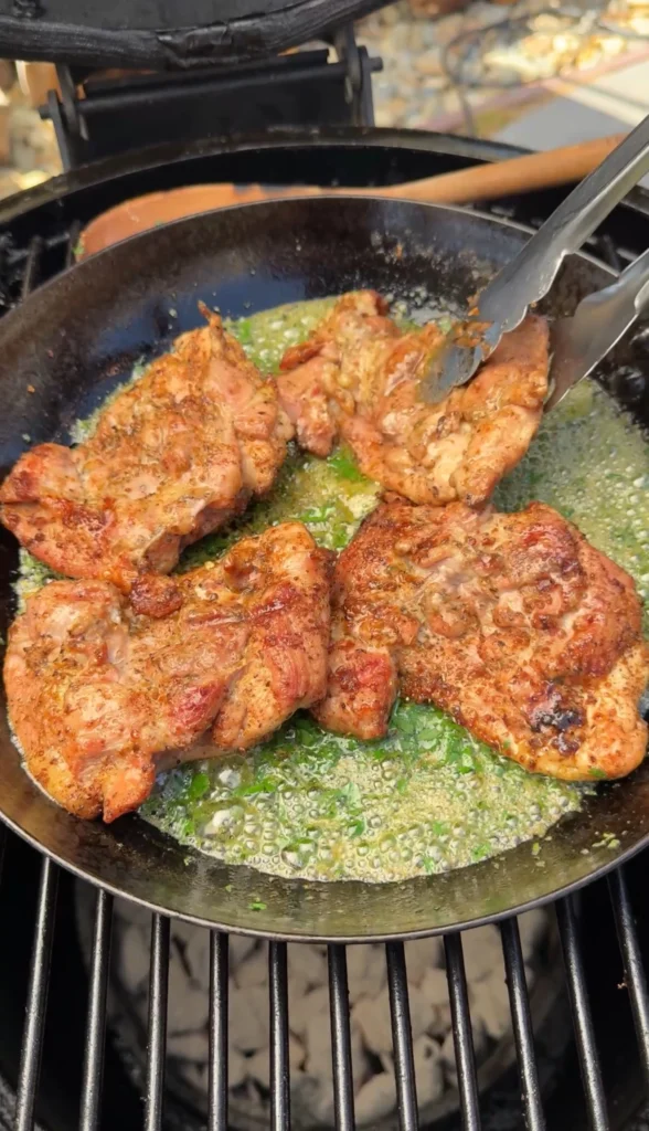 Chicken thighs cooking in cast iron skillet with melted garlic herb butter sauce, showing herbs and seasonings bubbling around golden chicken