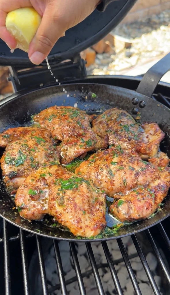 Grilled chicken thighs in cast iron skillet with garlic herb butter sauce, showing golden-brown color and fresh herb garnish on grill grates