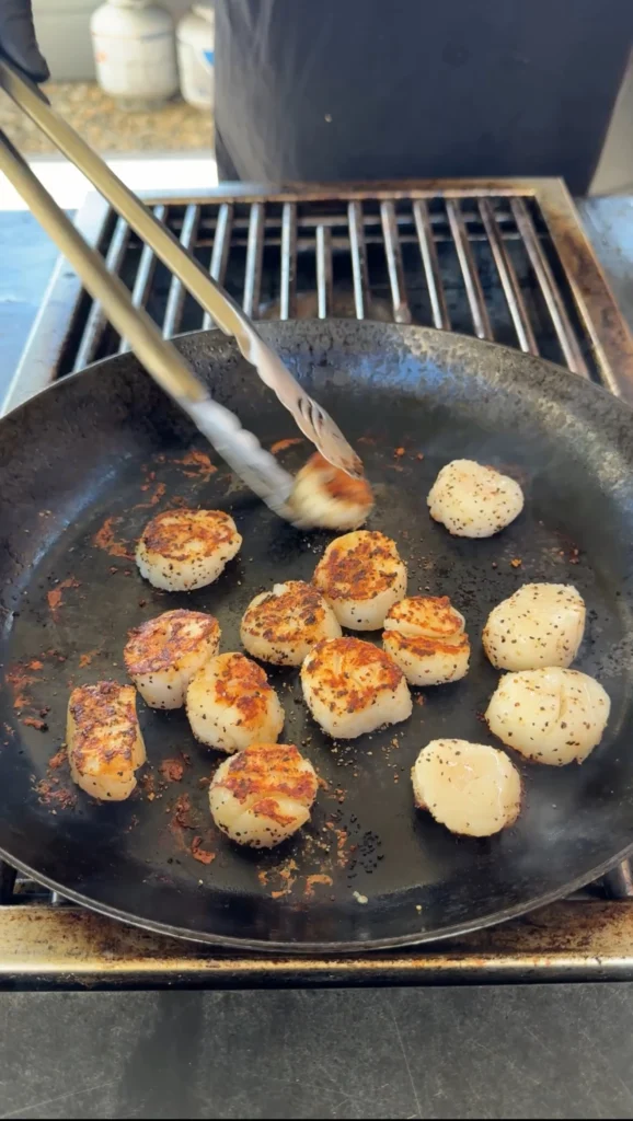 Jumbo scallops being turned with tongs in hot cast iron skillet, showing golden-brown searing and proper cooking technique