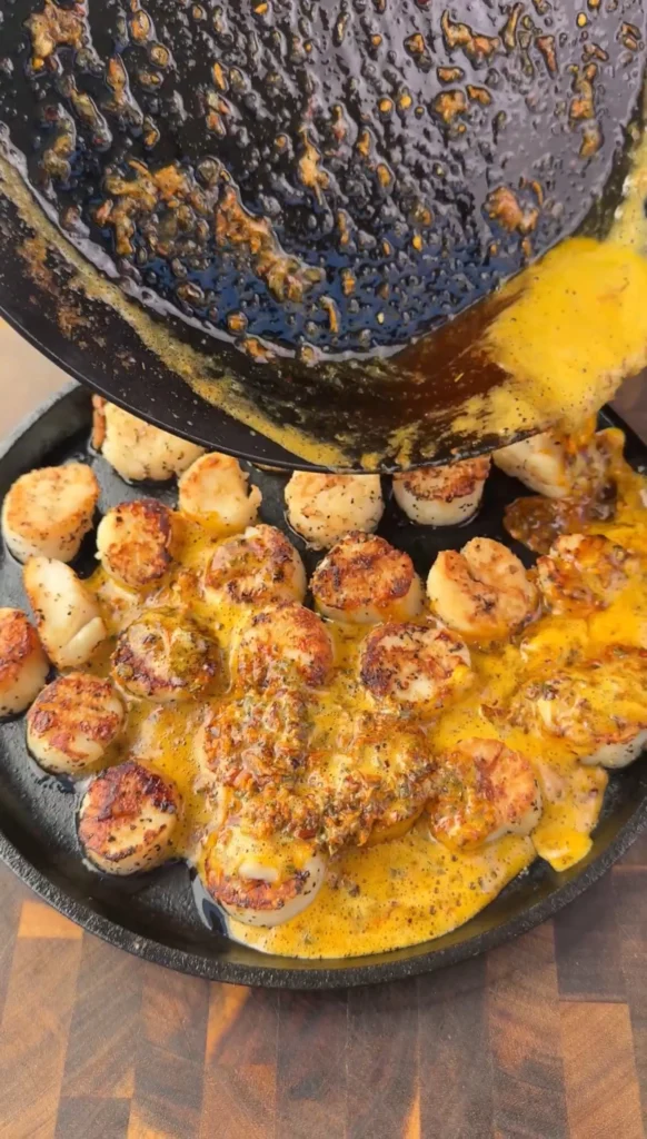 Seared scallops in cast iron pan with melted cowboy butter sauce bubbling around them, showing rich golden color and herb distribution
