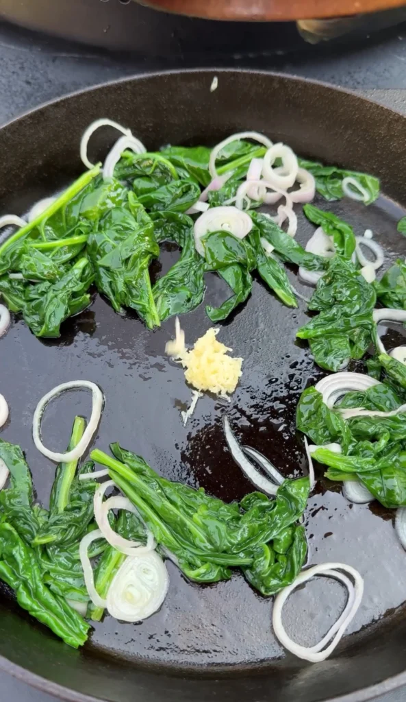 Fresh spinach and sliced shallots sautéing in a black skillet with minced garlic for chili oil steak pinwheel filling