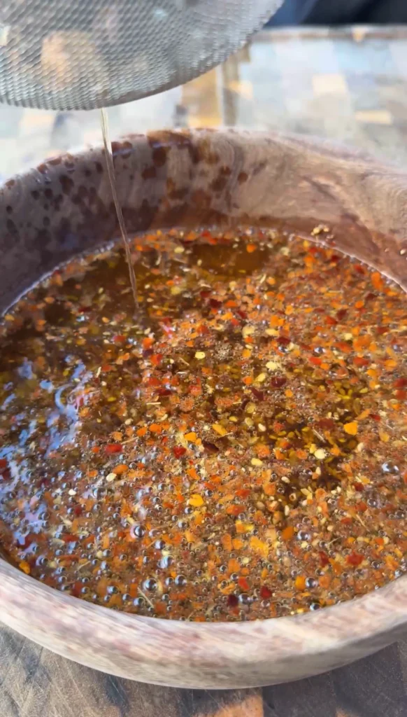 Close-up of bubbling homemade chili oil in a wooden bowl showing red pepper flakes, spices, and aromatic ingredients
