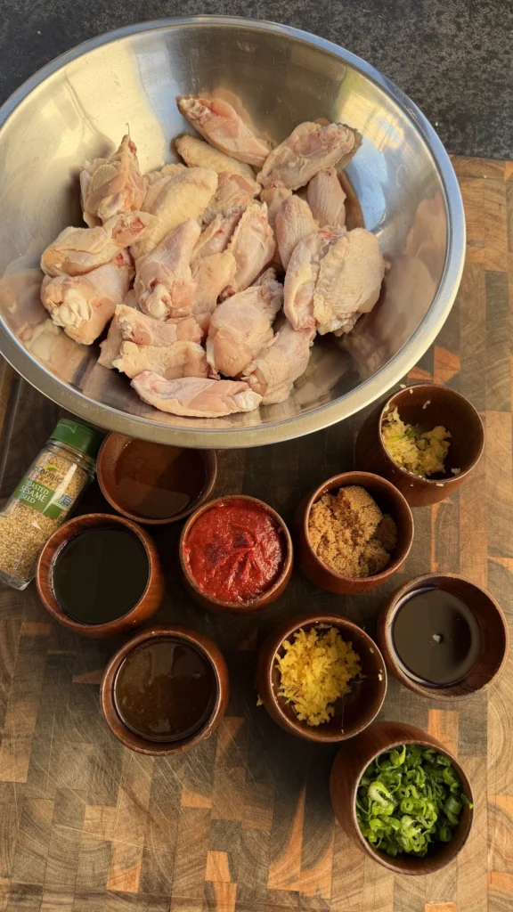 Raw chicken wings in a steel bowl with gochujang, brown sugar, garlic, ginger, soy sauce, and sesame seeds in wooden prep bowls on a cutting board