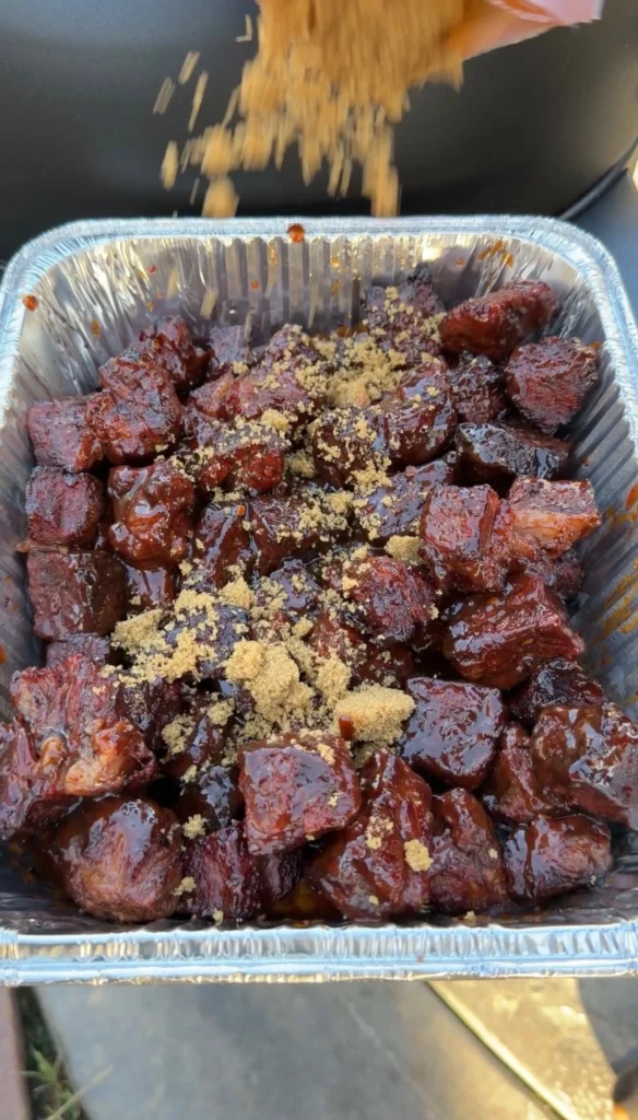 Brown sugar being sprinkled over sauced chuck roast burnt ends in a foil pan