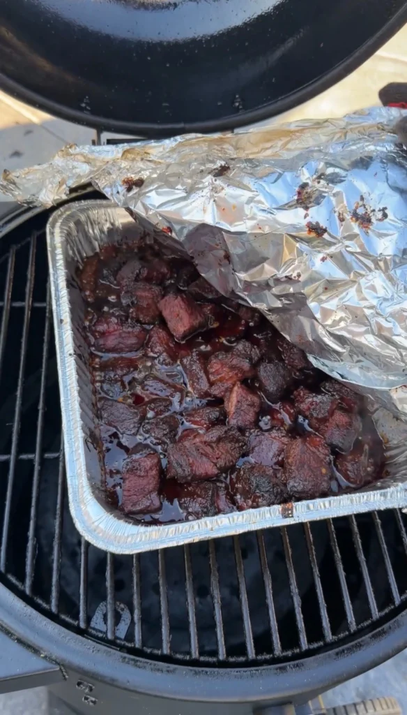 Chuck roast burnt ends braising in BBQ sauce in a foil-covered pan on the smoker