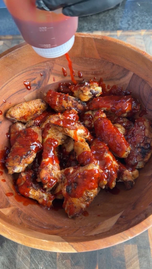 Gochujang sauce being poured from a squeeze bottle over grilled chicken wings in a wooden tossing bowl