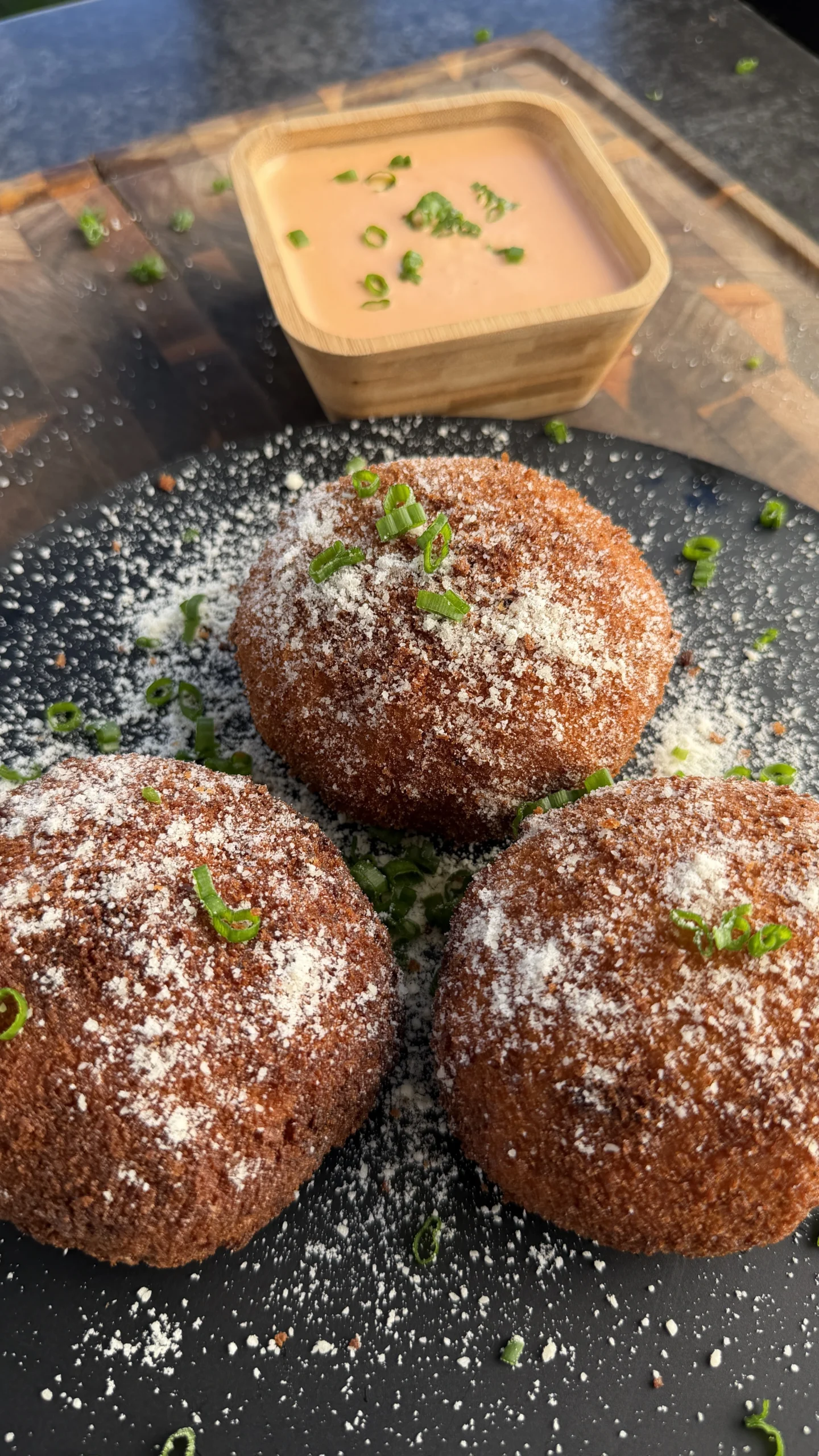 Three golden fried buffalo chicken potato croquettes on a dark plate with grated Parmesan, green onions, and ranch dipping sauce