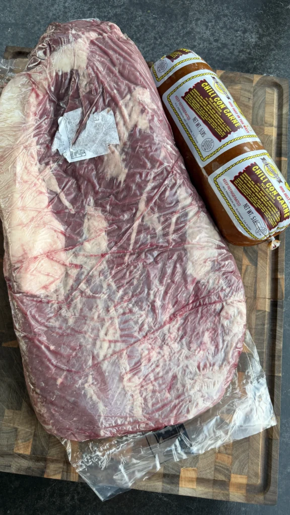Raw brisket flat in packaging with two RC Provisions chili con carne logs on a cutting board