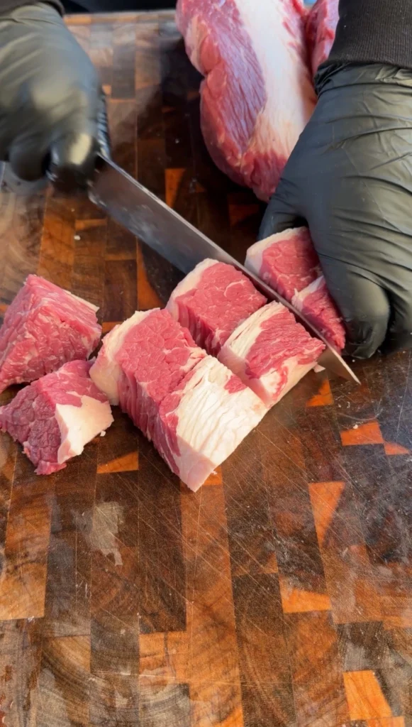 Hands in black gloves cutting raw brisket point into 1-inch cubes on a cutting board