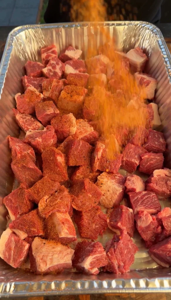 Cubed brisket point in a foil pan being generously coated with BBQ seasoning