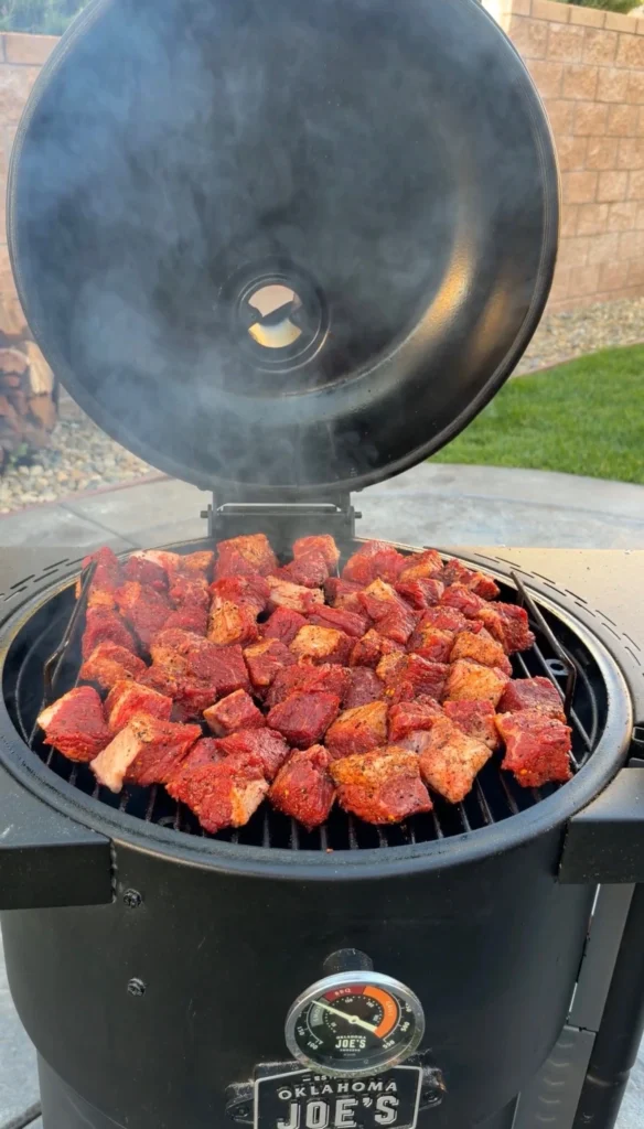 Seasoned brisket cubes smoking on an Oklahoma Joe's smoker with smoke billowing