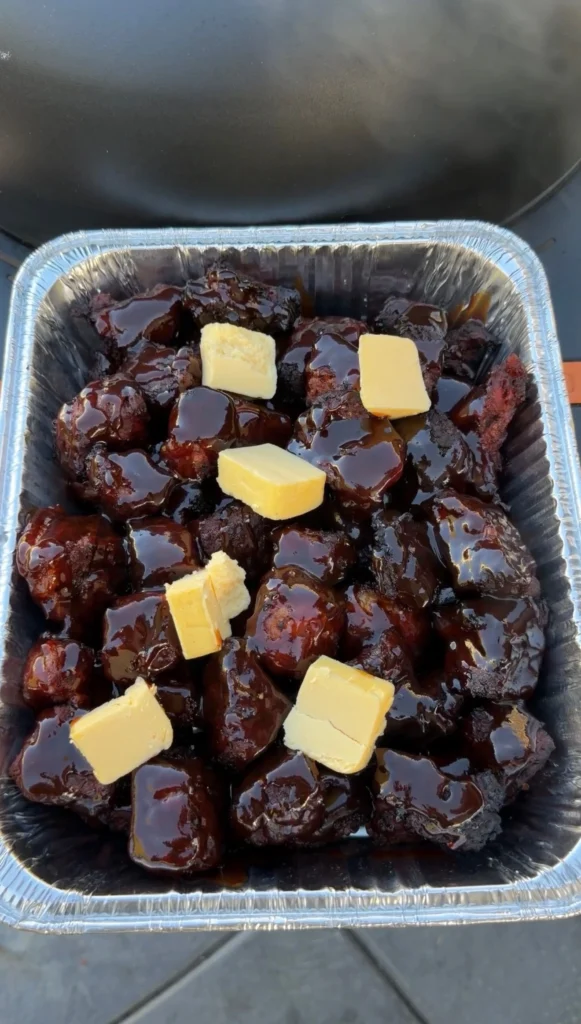 Brisket burnt ends in a foil pan coated in BBQ sauce with butter pats on top ready for braising