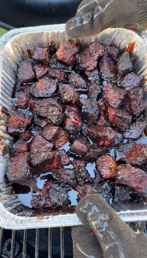 Finished smoked brisket burnt ends with sticky caramelized glaze held in a foil pan with gloved hands