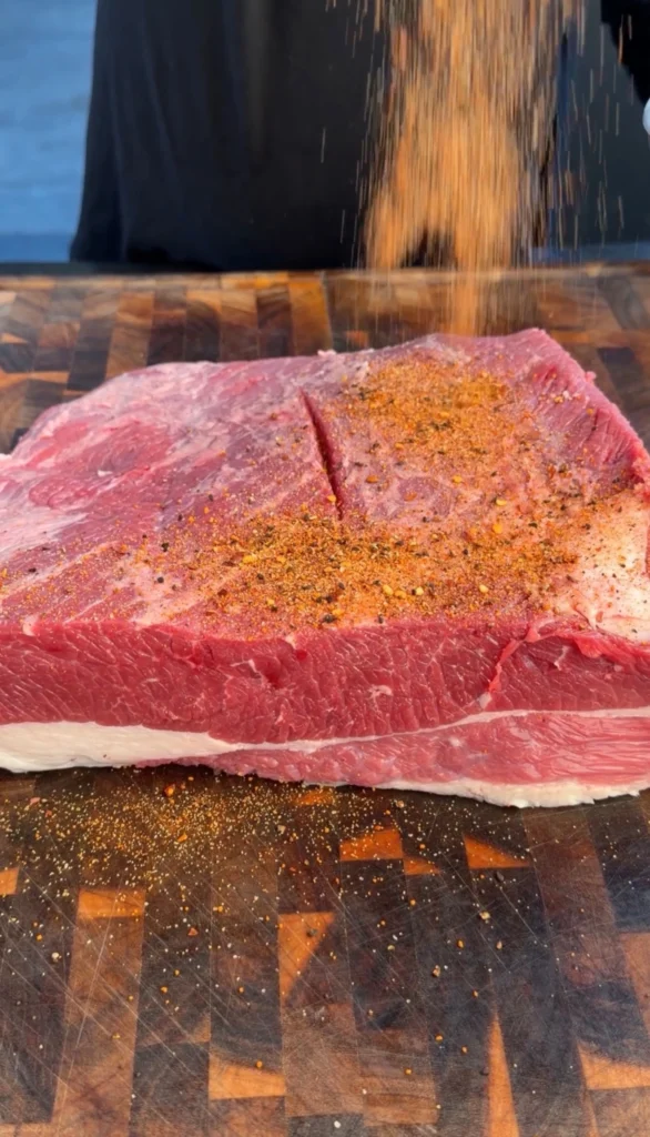 Raw brisket flat being seasoned with BBQ rub on a cutting board