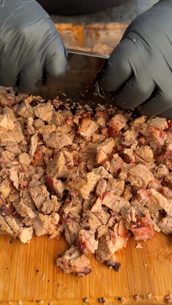 Hands in black gloves chopping smoked brisket into small chunks on a cutting board