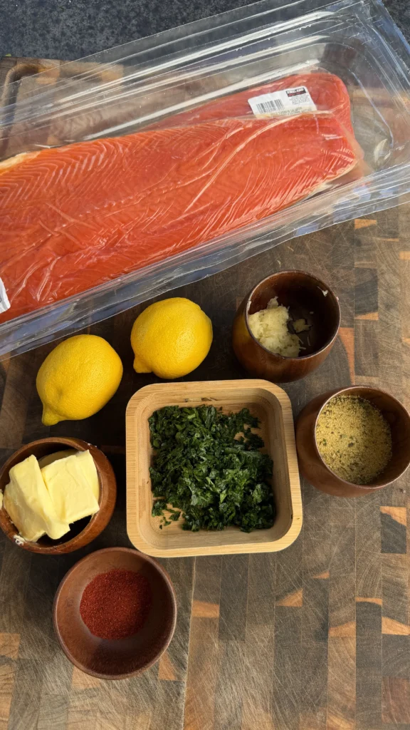 Raw salmon fillet with lemons, minced garlic, parsley, lemon pepper seasoning, paprika, and butter in wooden bowls on a cutting board