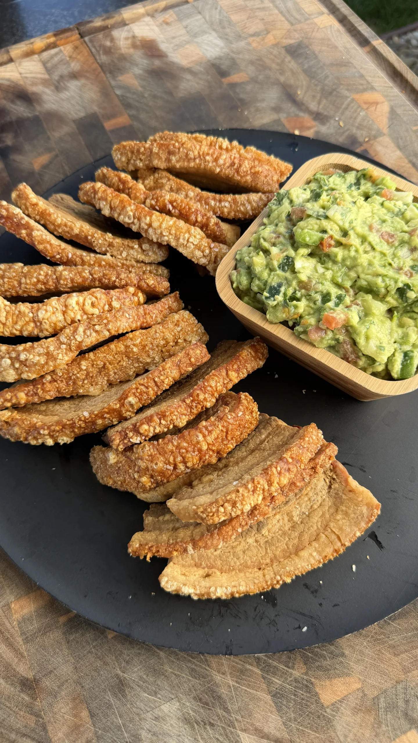 Crispy chicharrón chips sliced and fanned on a black plate with a wooden bowl of fresh guacamole