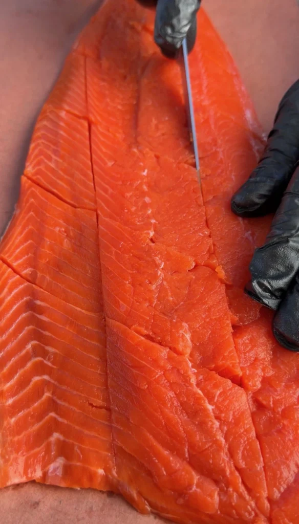 Close-up of hands in black gloves scoring a crosshatch pattern into a raw salmon fillet on butcher paper