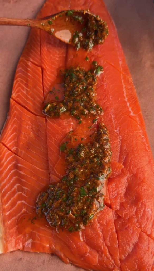 Wooden spoon spreading lemon pepper garlic herb butter mixture over a scored raw salmon fillet on butcher paper