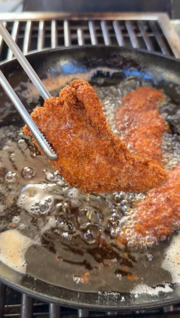 Golden brown crispy fried chicken tender lifted from oil with tongs showing crunchy panko crust