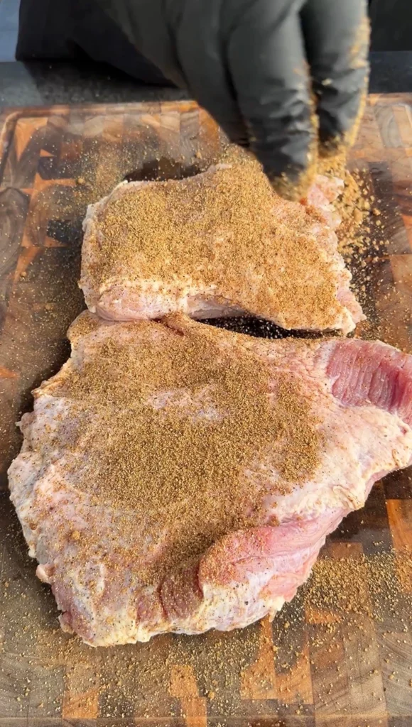 Two corned beef flats generously coated with pastrami rub on a cutting board with gloved hands