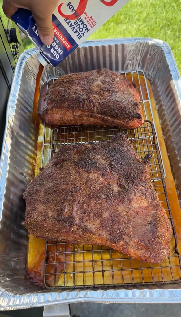 Smoked corned beef on a wire rack in a foil pan with beef broth being poured underneath for braising