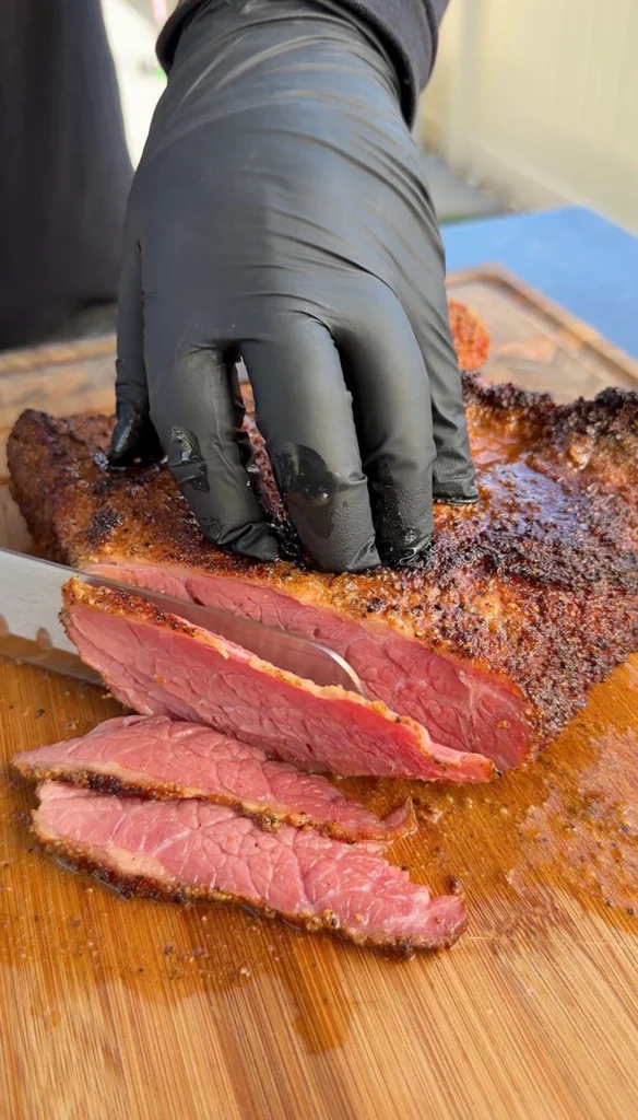 Hands in black gloves slicing smoked pastrami thin on a cutting board showing pink interior