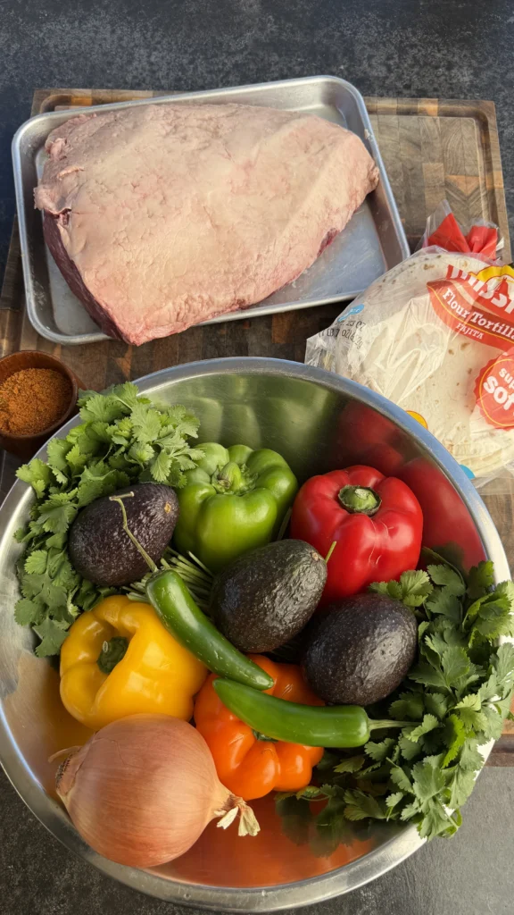 Raw picanha roast on a sheet pan with bell peppers, onion, avocados, cilantro, jalapeños, seasoning, and flour tortillas on a cutting board