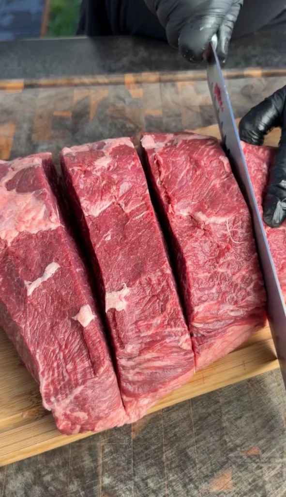 Hands in black gloves cutting a raw picanha roast into four thick steaks on a cutting board
