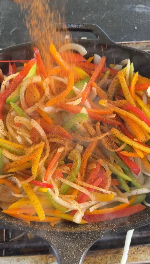 Sliced bell peppers and onions cooking in a cast iron skillet with fajita seasoning being sprinkled over them
