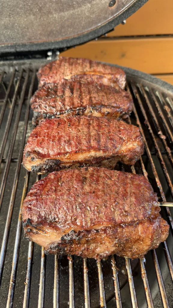 Four seared New York strip steaks with deep brown crust on kamado grill grates
