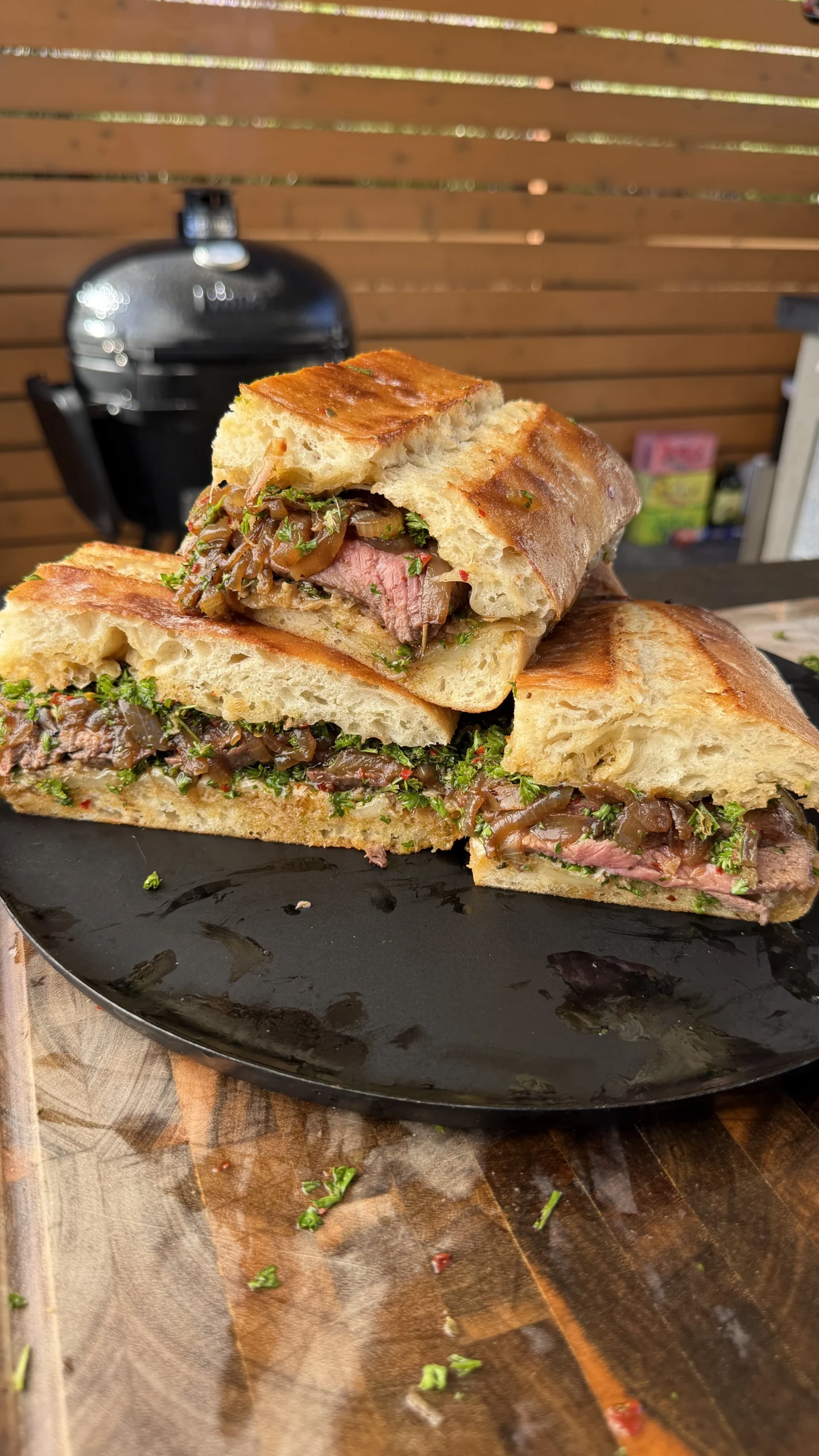 Sliced loaded steak sandwich on ciabatta with chimichurri, caramelized onions, and melted mozzarella on a black plate with kamado grill in background