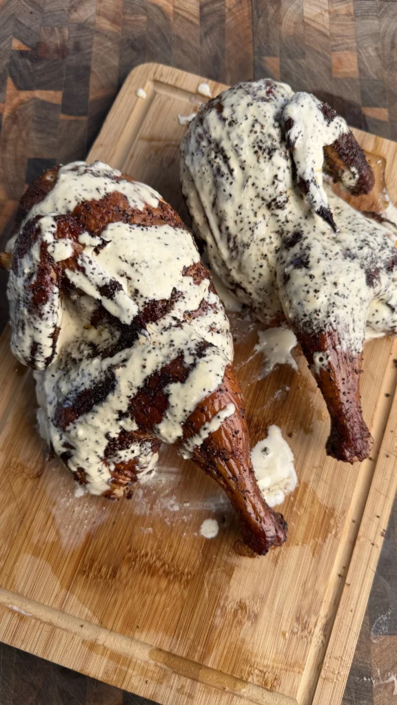 Two applewood smoked half chickens with creamy white sauce and black pepper on a wooden cutting board