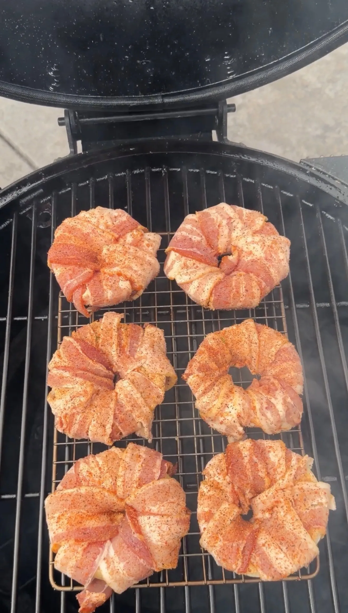 Six bacon-wrapped onion rings with seasoning cooking on a round BBQ grill grate