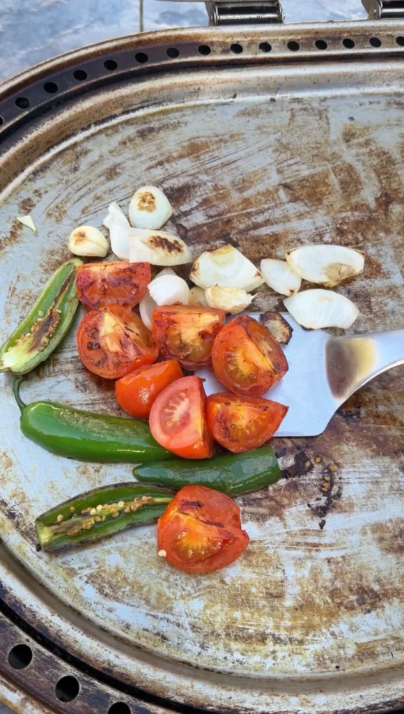 Charred tomatoes, peppers, and onions grilling on a metal comal for steak taco salsa