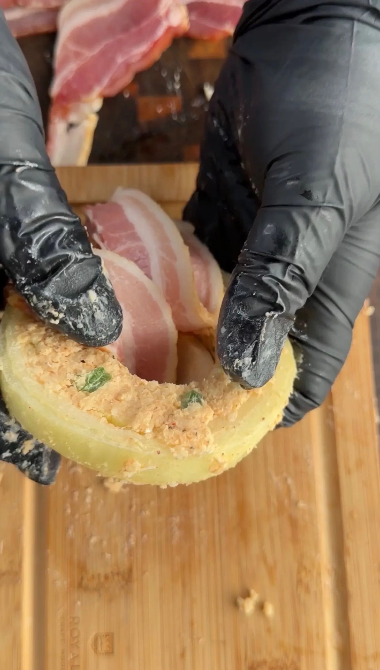 Hands in black gloves preparing crab stuffed bacon-wrapped onion rings on a wooden cutting board