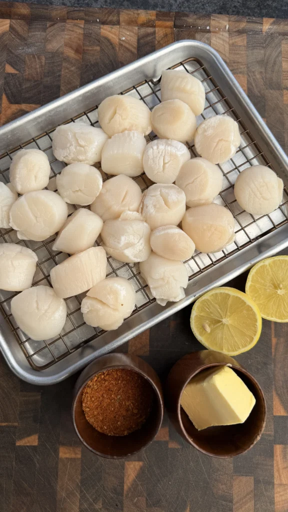 Raw sea scallops arranged on wire cooling rack with lemon halves, butter, and spice bowls on wooden board