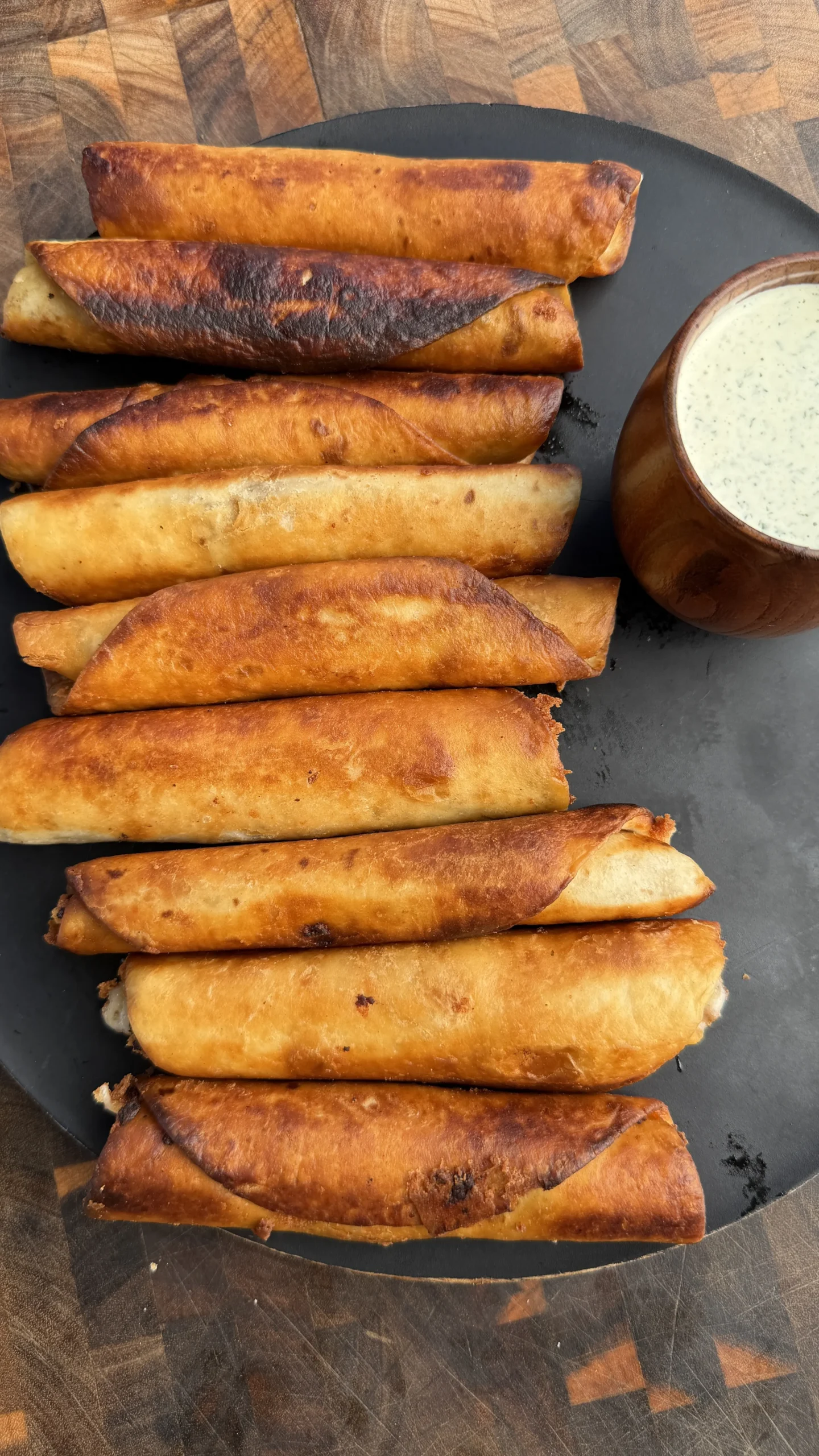 Golden brown crispy beef taquitos stacked on dark plate with white creamy dipping sauce in wooden bowl