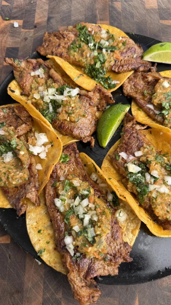 Grilled steak tacos with charred salsa, diced onions, and cilantro on corn tortillas served on a black slate plate
