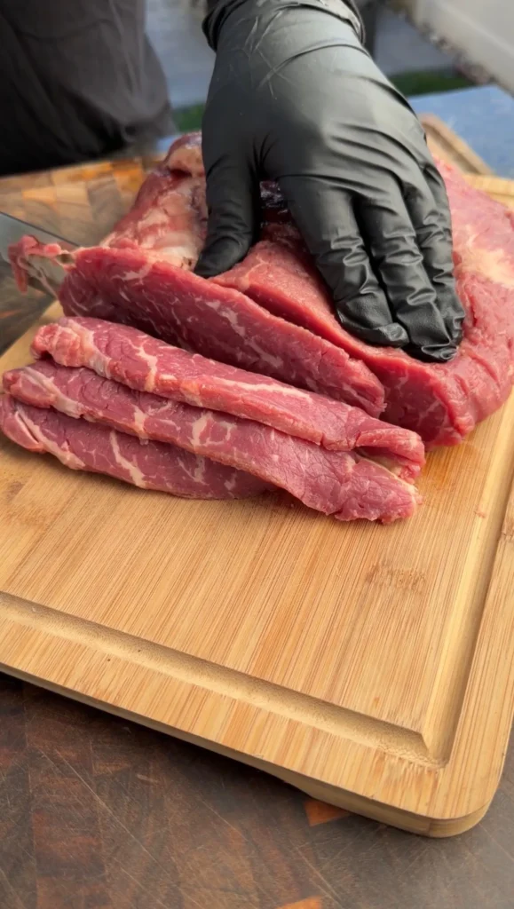 Raw beef strips being cut on wooden cutting board with gloved hands for steak tacos preparation