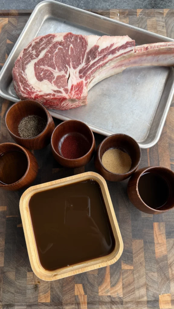Raw cowboy ribeye steak on metal tray with spice bowls and dark worcestershire glaze in wooden container
