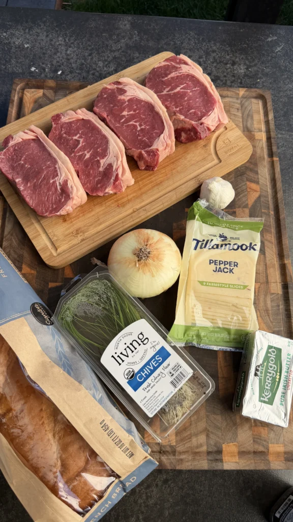 Raw ribeye steaks on cutting board with onion, chives, pepper jack cheese and bread for steak stuffed bread boat recipe