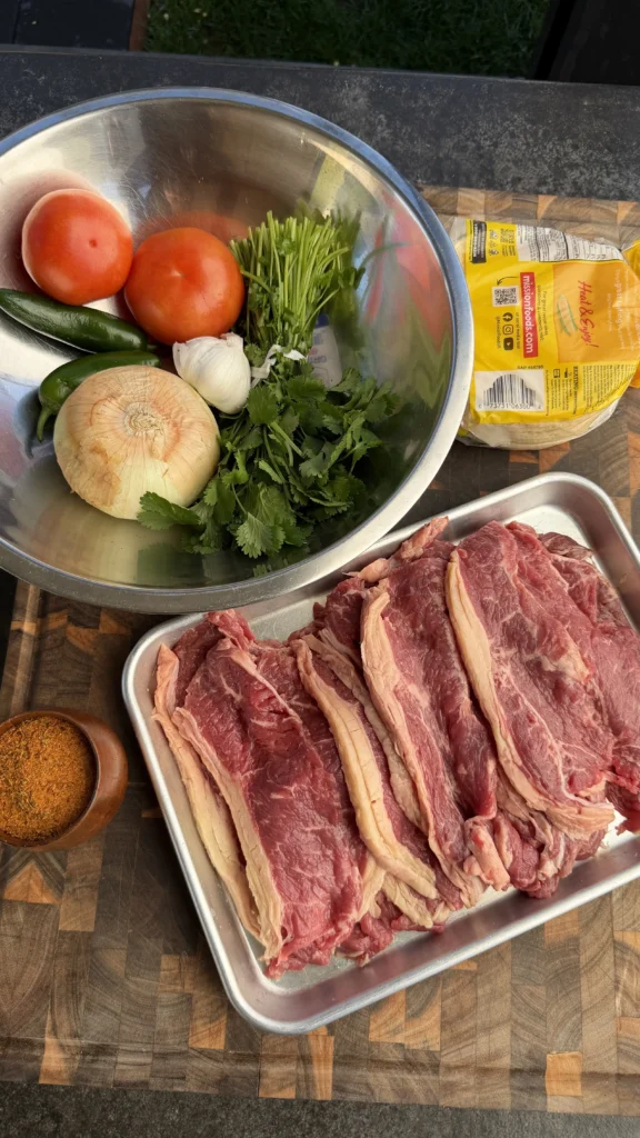 Raw beef strips in metal tray with fresh tomatoes, onion, cilantro, and tortillas for steak tacos