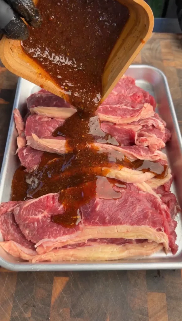 Raw beef steaks being coated with dark marinade sauce from wooden spoon in metal tray for steak tacos