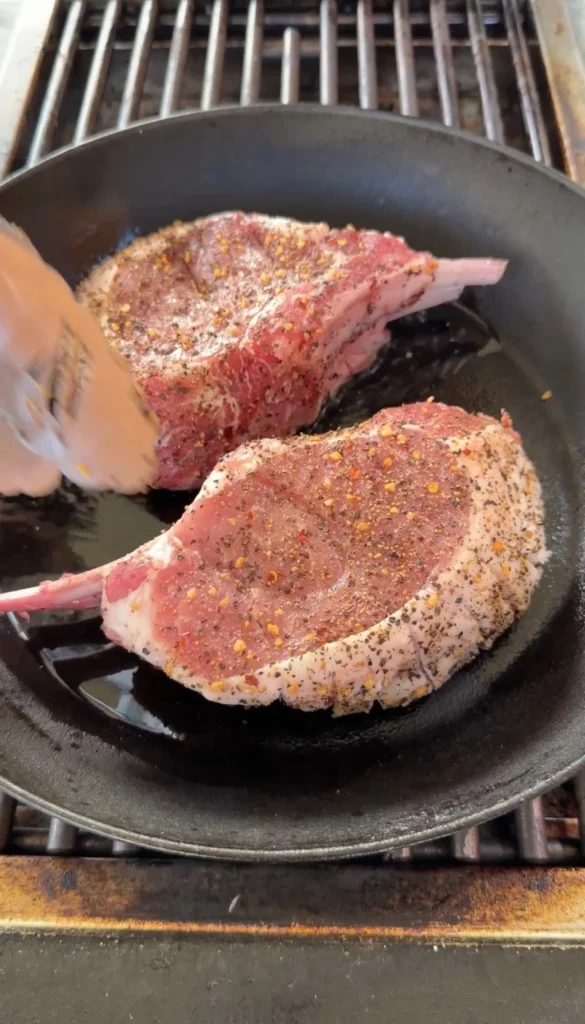 Two thick double bone pork chops seasoned with herbs and spices searing in a black cast iron skillet